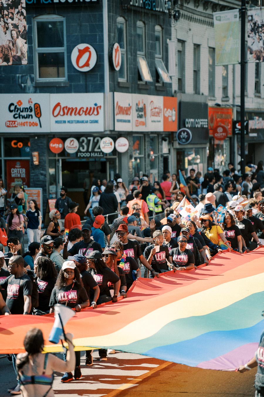 Participants in a parade carry a large rainbow flag, symbolizing LGBTQ+ pride, as they march past storefronts and spectators.