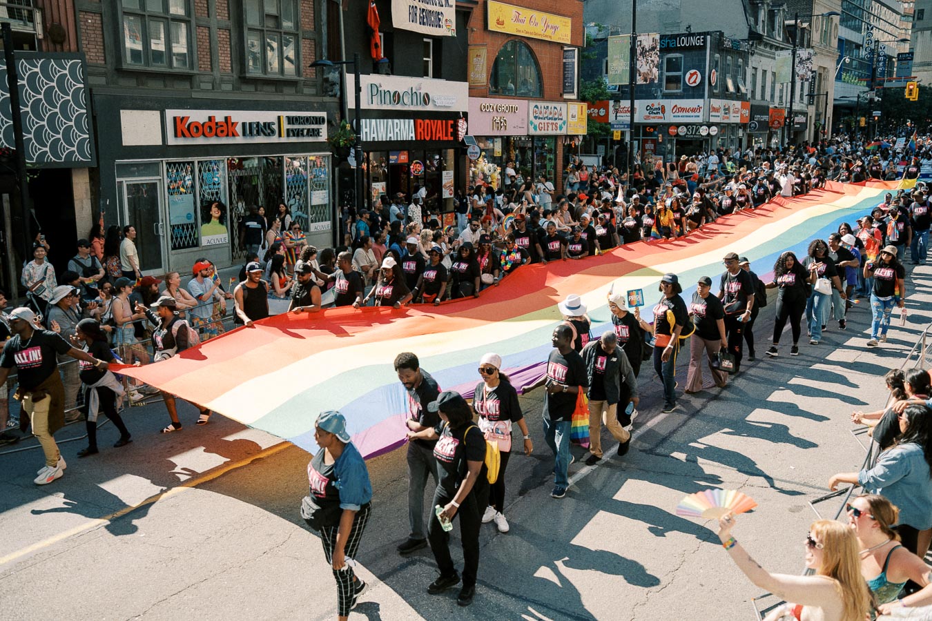Participants in a pride parade carry a large rainbow flag down a bustling city street, surrounded by cheering spectators.