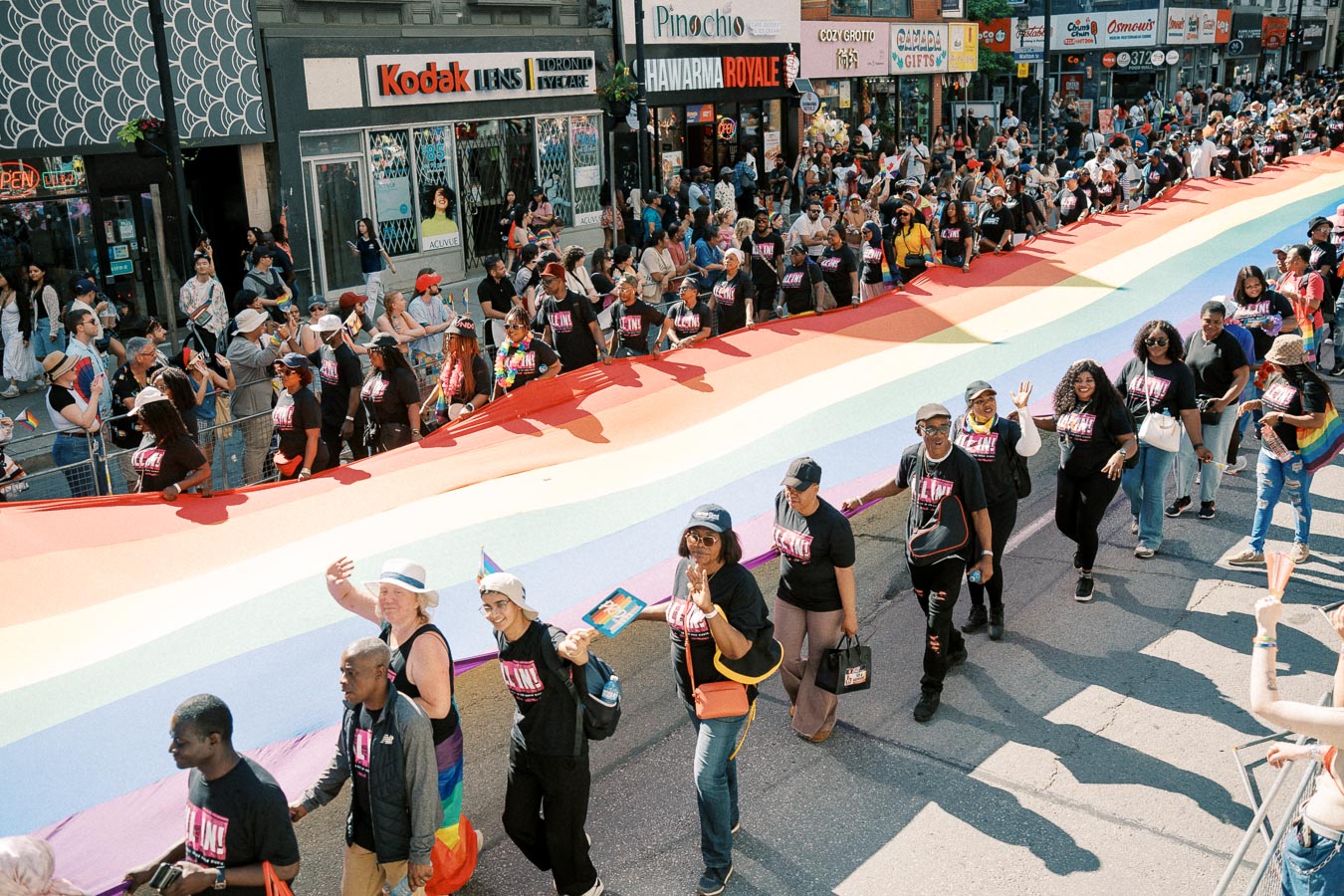 Participants march with a giant rainbow flag during a pride parade, surrounded by spectators.
