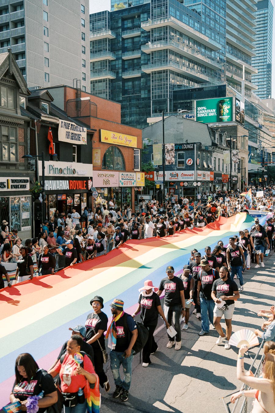 Participants in a Pride parade carry a large rainbow flag through a bustling city street, with onlookers lining the sidewalks.