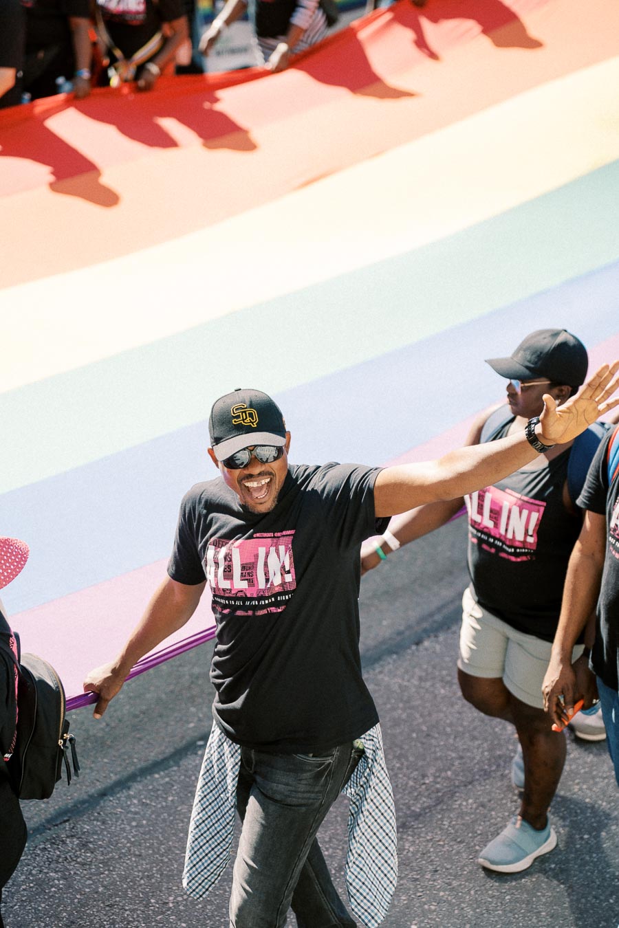 A joyful participant waves during a parade, walking alongside a large rainbow flag, symbolizing LGBTQ+ pride.
