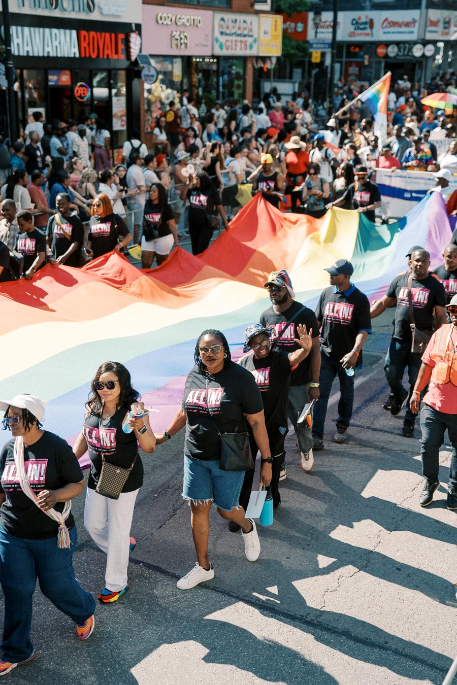 Participants in a pride parade march down the street, holding a large rainbow flag. They are wearing matching black shirts with "ALL IN!" printed on them. The street is lined with spectators and various shops.