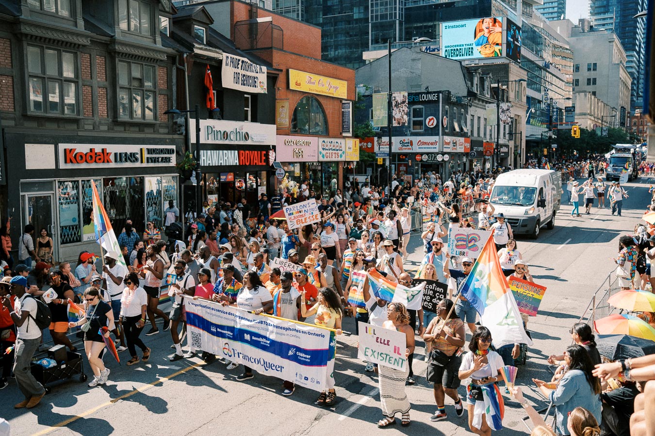 A lively LGBTQ+ pride parade takes place on a bustling city street, with participants carrying rainbow flags and banners. The street is lined with spectators and various shops, creating a festive atmosphere.