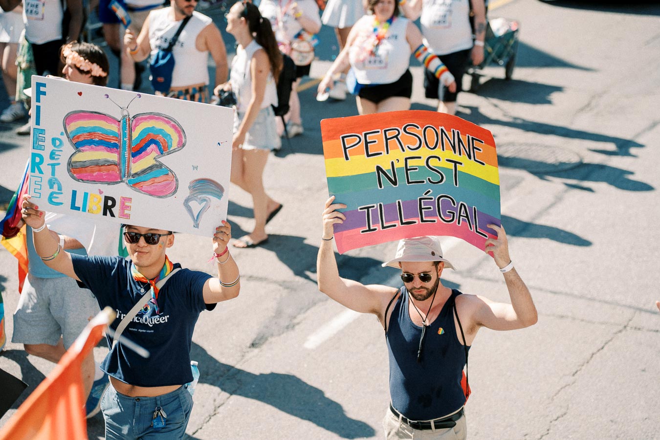 Participants at a pride parade hold colorful signs promoting freedom and inclusivity, with messages in French supporting LGBTQ+ rights and immigration equality.
