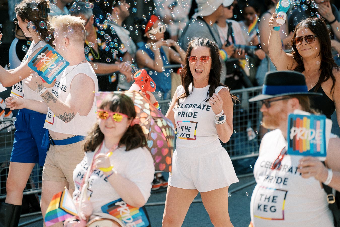 Participants joyfully celebrate at a Pride parade, wearing themed shirts and holding rainbow flags, with bubbles floating around.