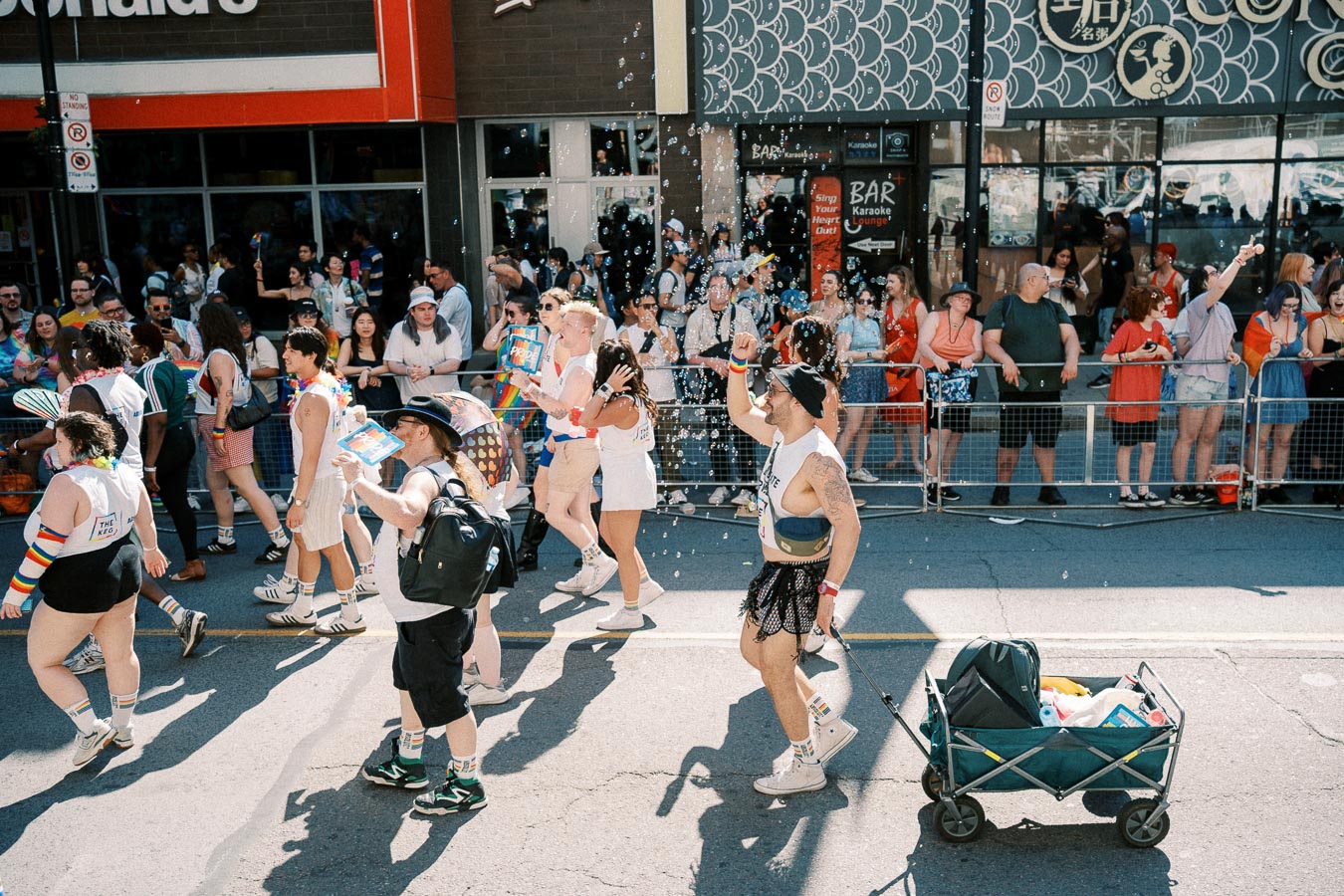 Participants in a vibrant parade march down the street, adorned in colorful attire and rainbow-themed accessories, while spectators watch from the sidelines. Bubbles add to the festive atmosphere.