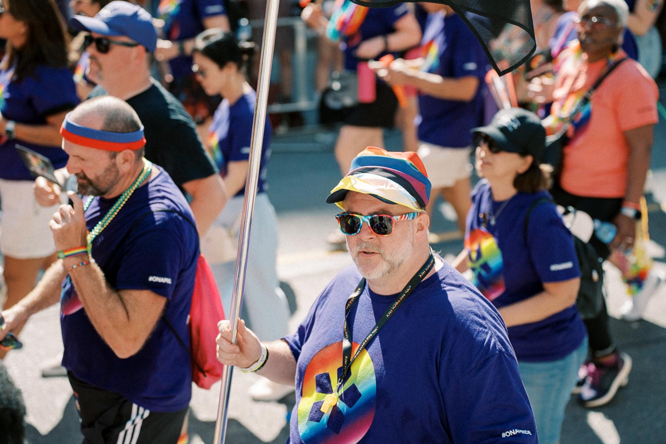 Participants in a pride parade wearing purple shirts with rainbow designs and colorful accessories.