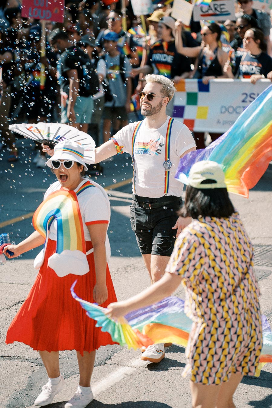 Participants at a pride parade joyfully celebrate with rainbow-themed outfits and accessories, surrounded by bubbles.