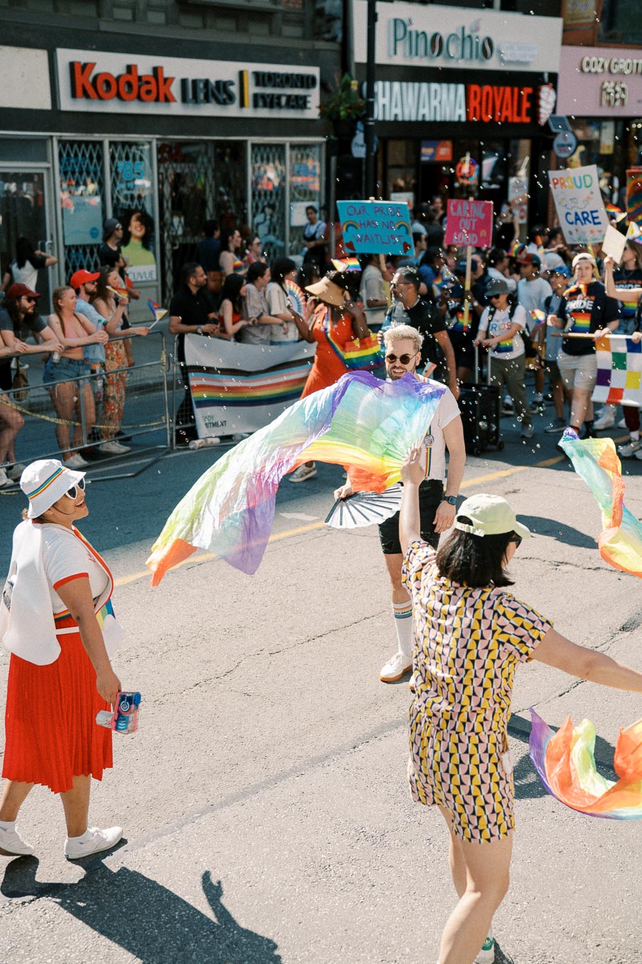Participants joyfully celebrate at a pride parade, waving rainbow flags and wearing vibrant outfits. The street is lined with spectators, and shops are visible in the background.