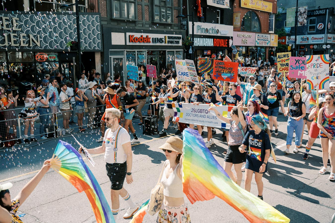 Participants in a lively Pride parade march down a city street, holding rainbow flags and signs promoting LGBTQ+ rights and healthcare equity.