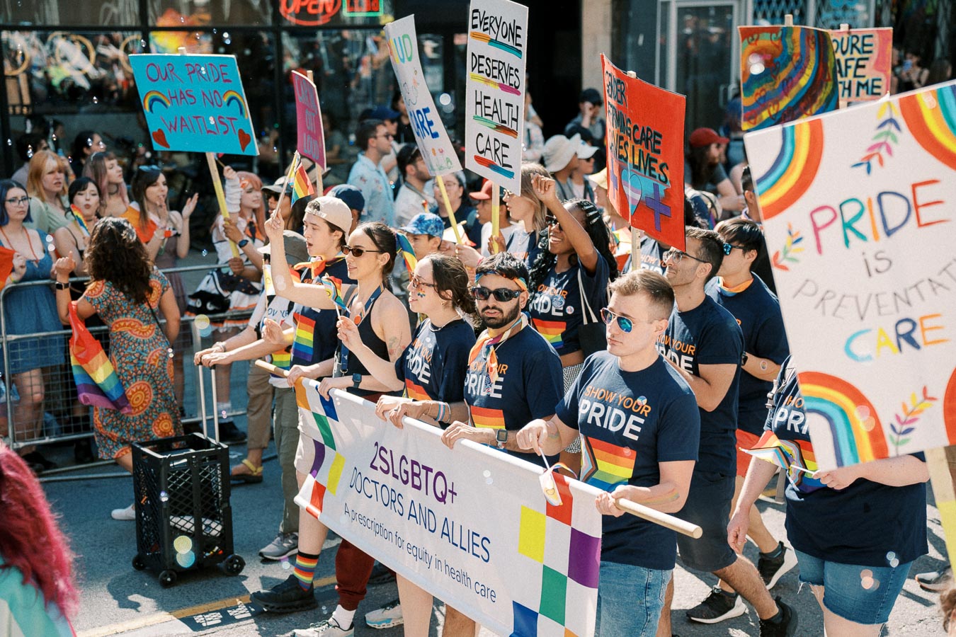 Participants in a Pride parade march with signs advocating for healthcare equality and LGBTQ+ rights, holding a banner for '2SLGBTQ+ Doctors and Allies'.