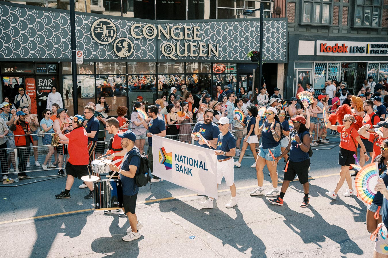 Participants in a parade hold a National Bank banner and rainbow fans, celebrating diversity and inclusion. Spectators line the sidewalk, enjoying the festive atmosphere.