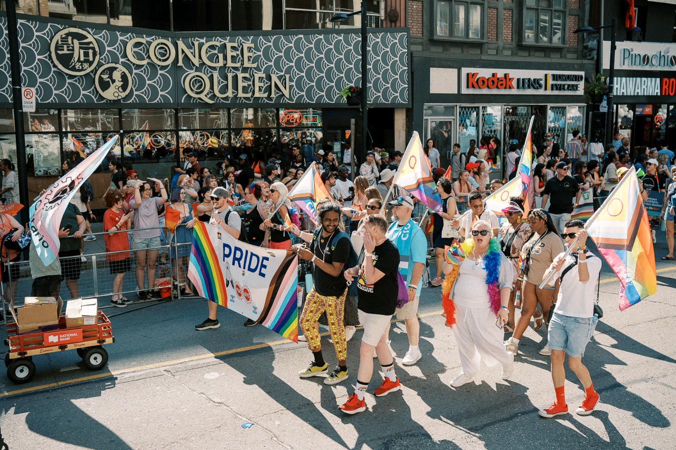 Participants march in a Pride parade, holding flags and a banner, with spectators lining the street.