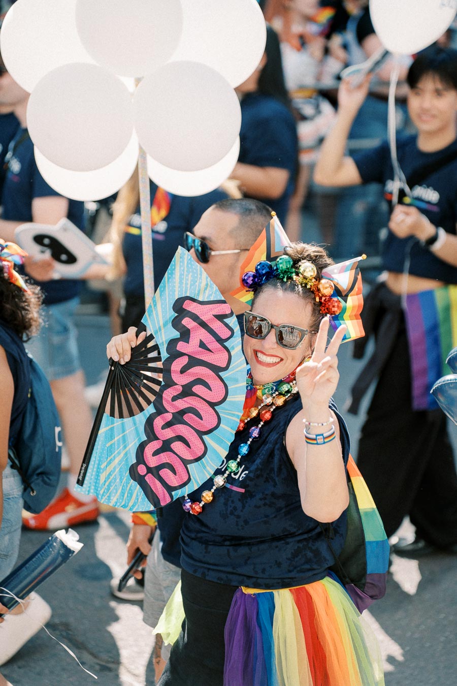 A joyful participant at a pride parade, adorned with rainbow-themed accessories and holding a "Yasss" fan, celebrates with white balloons.