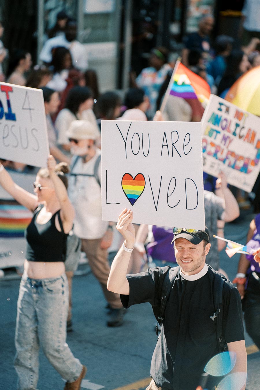 A participant in a pride parade holds a sign reading 'You Are Loved' with a rainbow heart, symbolizing support and inclusivity.