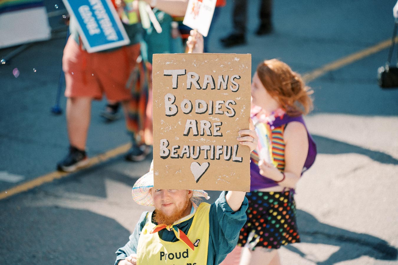 A person participates in a parade, holding a sign that reads 'Trans Bodies Are Beautiful,' promoting transgender pride and acceptance.