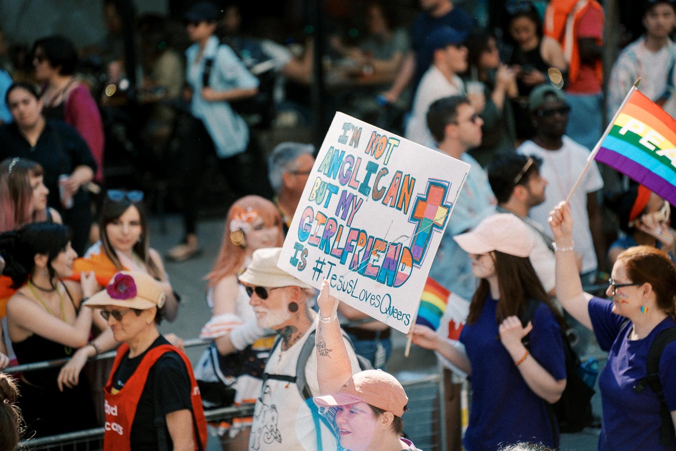 A vibrant scene from a pride parade featuring a person holding a sign that reads, 'I'm not Anglican but my girlfriend is #JesusLovesQueers'. The crowd is diverse, with rainbow flags and colorful attire.