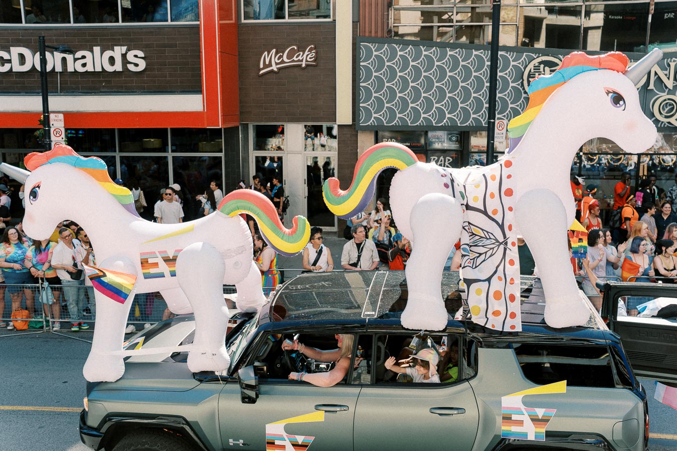 A vibrant parade float with inflatable unicorns and rainbow decorations passes by a McDonald's during a festive event.