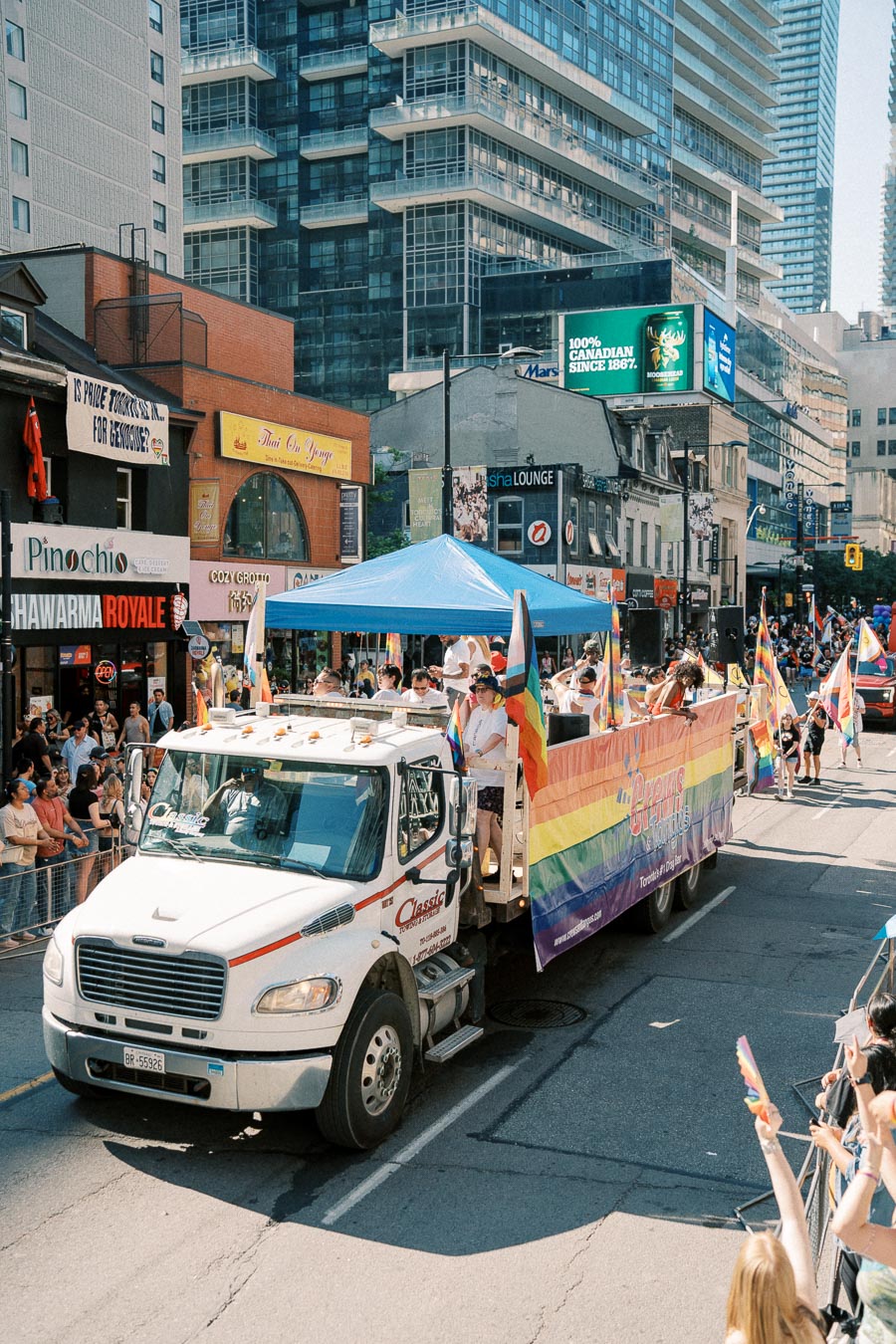 A vibrant parade float adorned with rainbow flags and banners moves through a city street, surrounded by cheering spectators. The event appears to be a pride parade, celebrating diversity and inclusion.