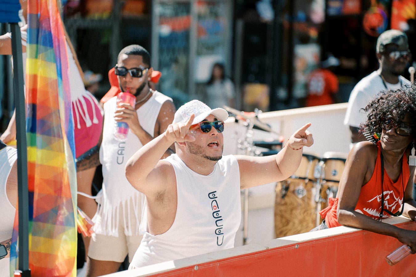 A lively scene at a parade featuring individuals in festive attire, with a prominent rainbow flag and musical instruments in the background, suggesting a celebration of diversity and inclusion.