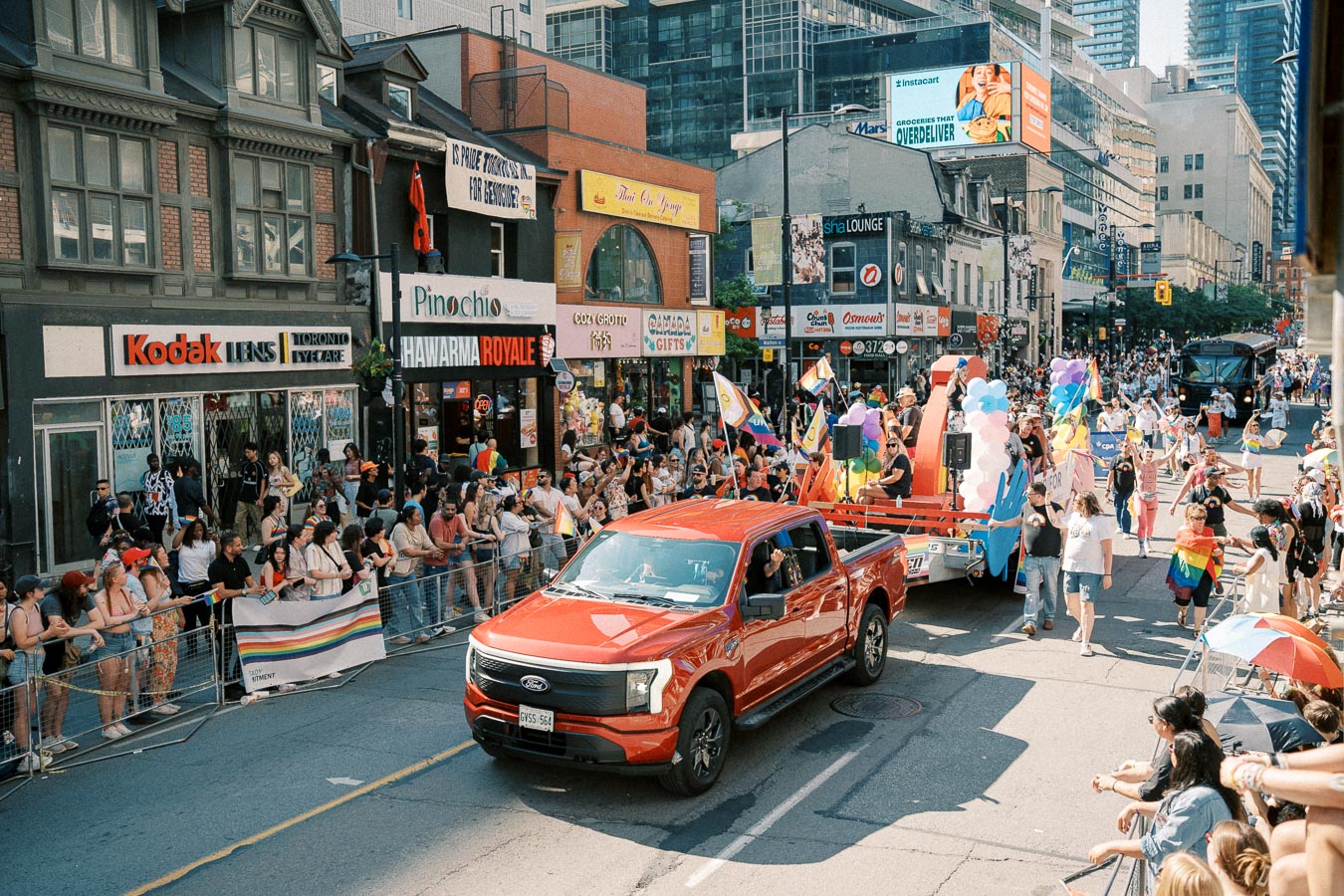 A lively pride parade featuring a red Ford truck and a colorful float, with spectators lining the street.