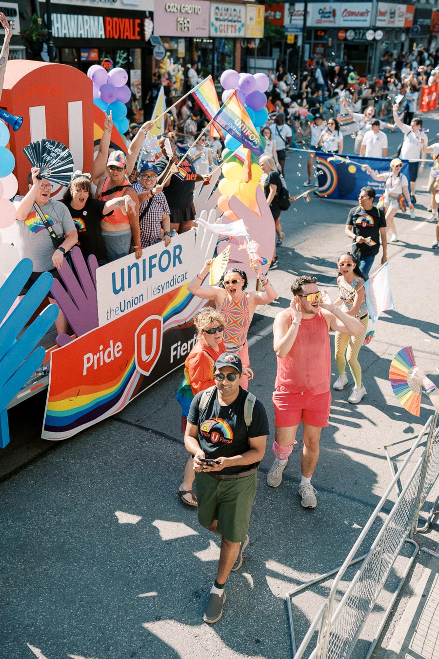 Participants celebrate at a Pride parade, showcasing colorful outfits and rainbow flags. A float with the Unifor logo and Pride banners adds to the festive atmosphere.