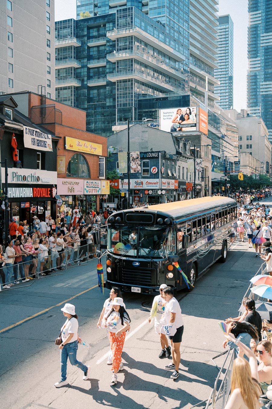 A vibrant street parade with a black bus and participants in colorful outfits, surrounded by a cheering crowd and urban buildings.