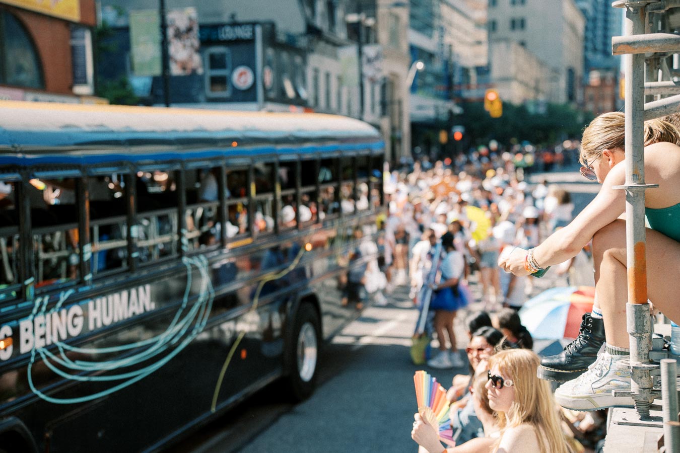 A person sits on a railing, observing a lively parade with a bus and a large crowd in the background, celebrating in a vibrant urban setting.