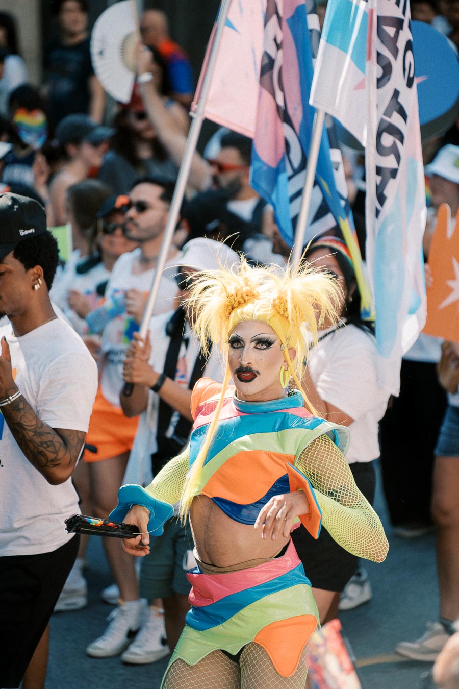 A vibrant scene at a pride parade featuring a person in a colorful drag costume with bright yellow hair, surrounded by people holding flags and fans.