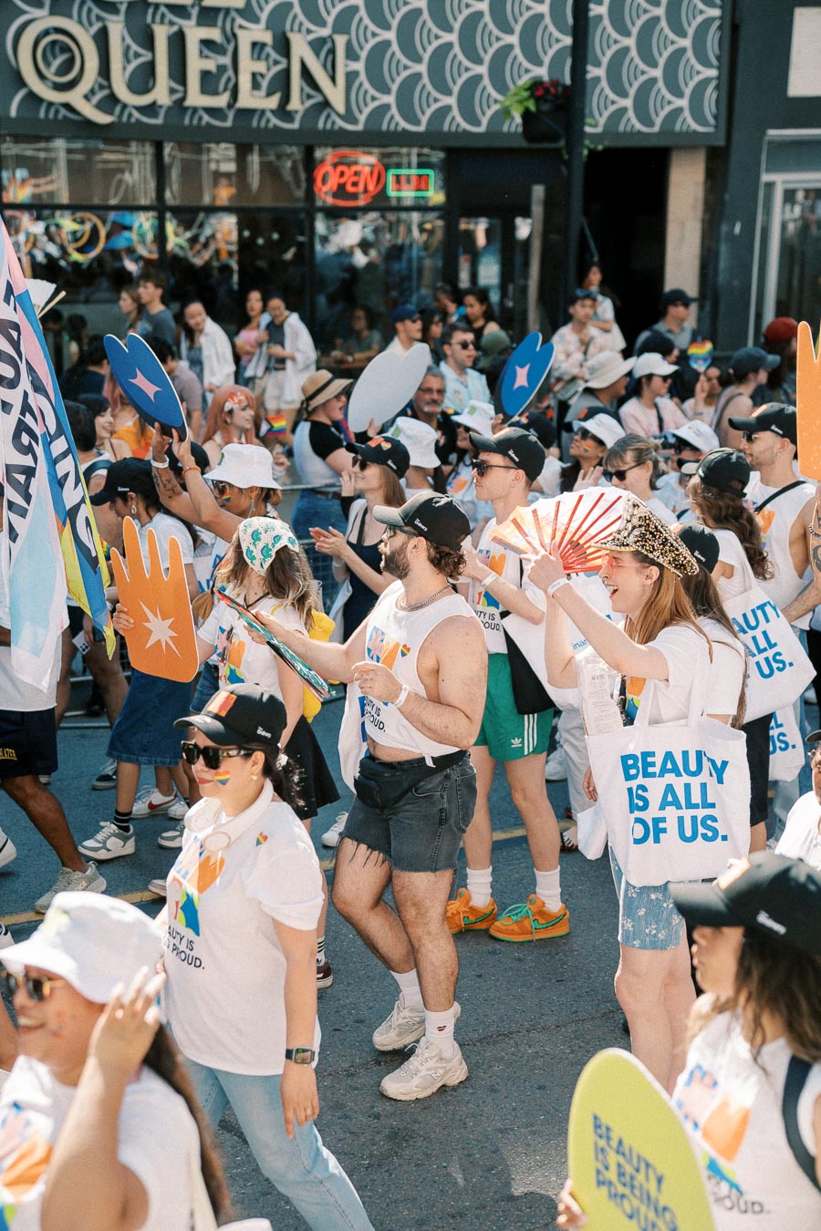 Participants in a pride parade celebrate with colorful attire and signs, marching past a storefront labeled 'QUEEN'.