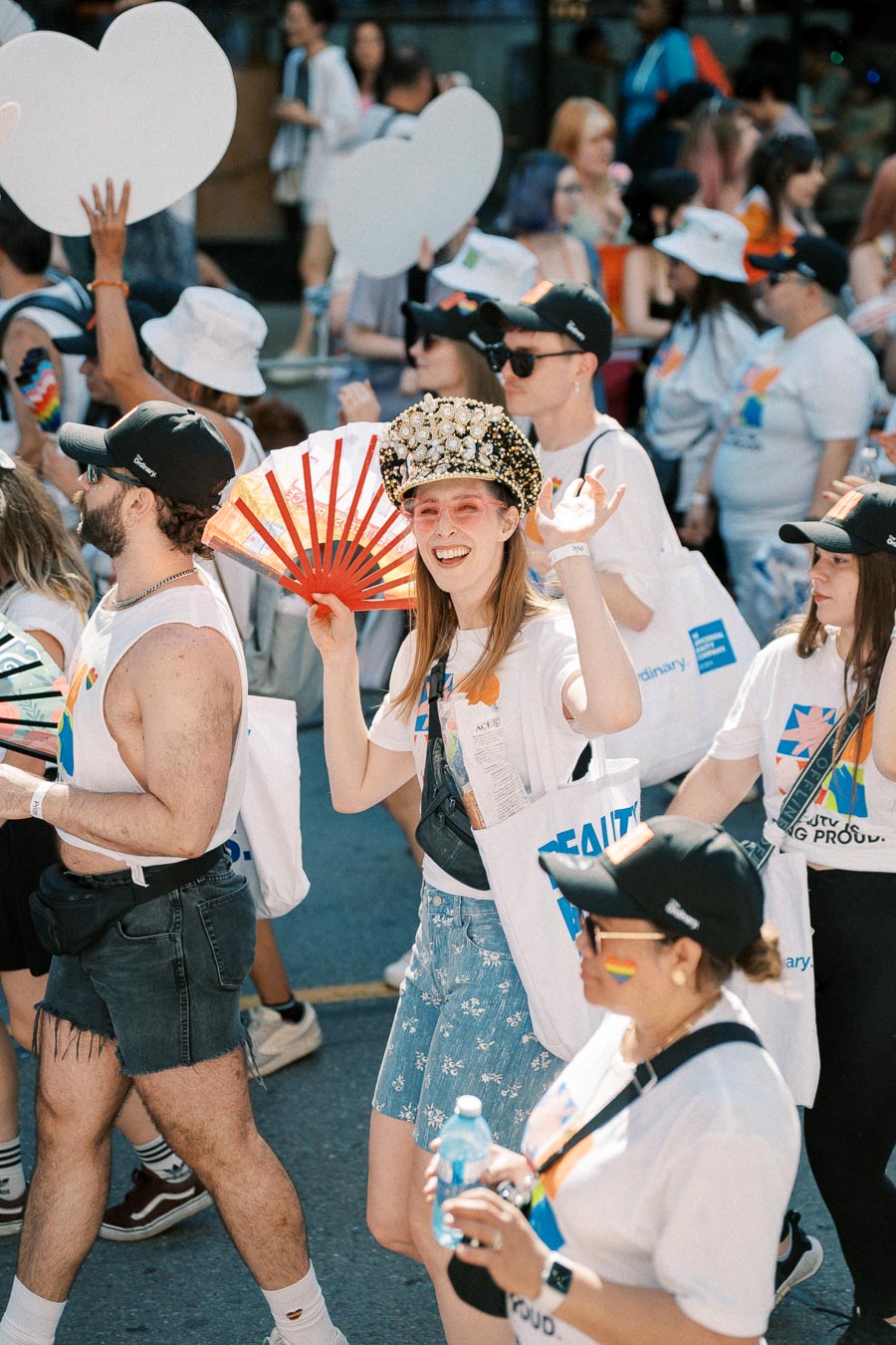 Participants in a pride parade celebrate with colorful outfits and accessories, including a red fan and a decorative hat.