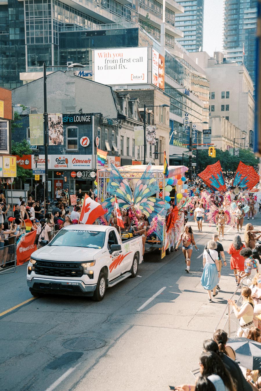 A lively parade scene featuring a white truck pulling a colorful float with participants in elaborate costumes, set against an urban backdrop with spectators and city buildings.