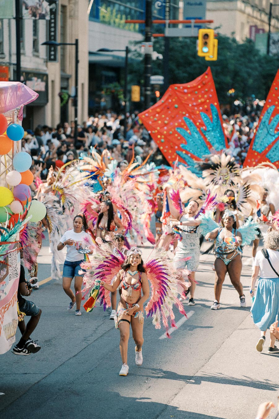 Participants in colorful feathered costumes parade through a city street during a lively festival, with a large crowd of spectators.