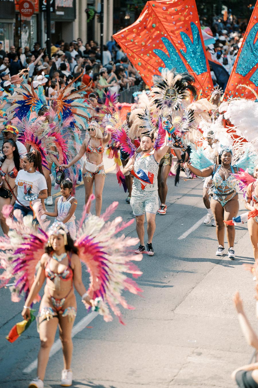 Participants in colorful feathered costumes dance energetically during a lively street parade, surrounded by a cheering crowd.