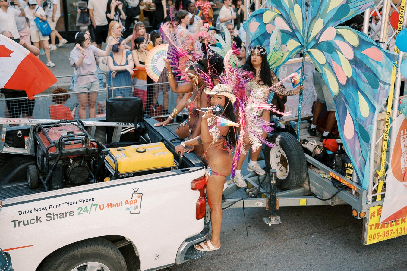 A lively parade featuring a woman in a colorful feathered costume on a U-Haul truck, celebrating with a rainbow fan and horn, amidst a cheering crowd and a Canadian flag.