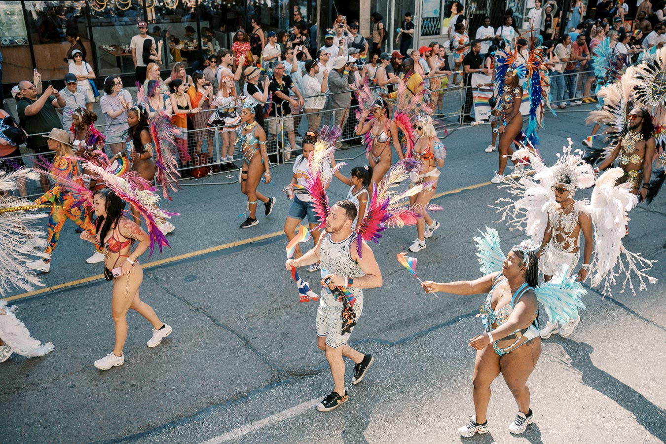 Participants in colorful costumes and feathered headdresses march in a lively parade, waving rainbow flags, as spectators watch from behind barriers.