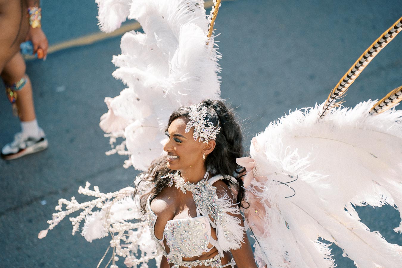 A woman dressed in an elaborate carnival costume featuring large white feathered wings and a sparkling headpiece participates in a parade.