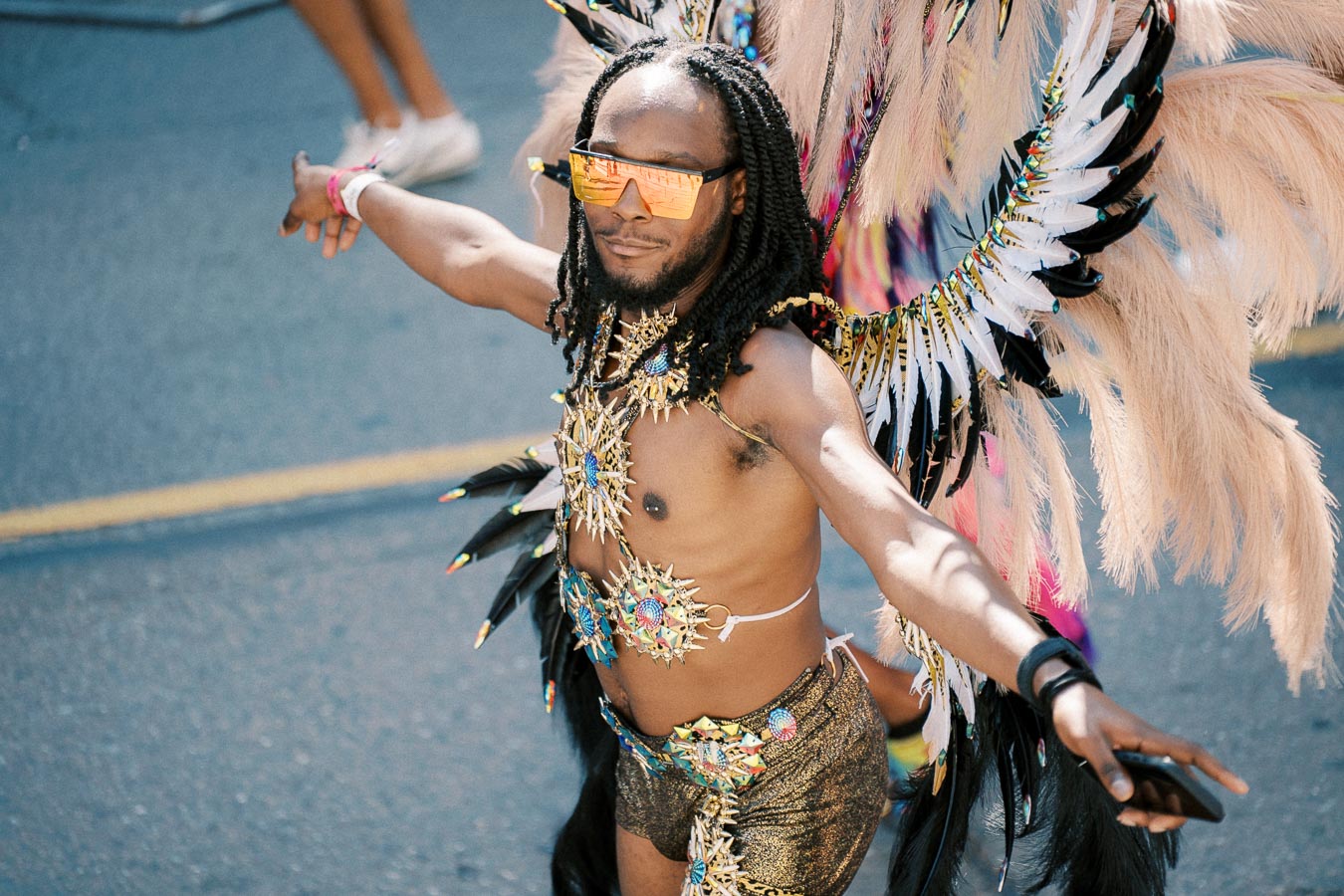 A vibrant carnival participant in an elaborate feathered costume and reflective sunglasses.