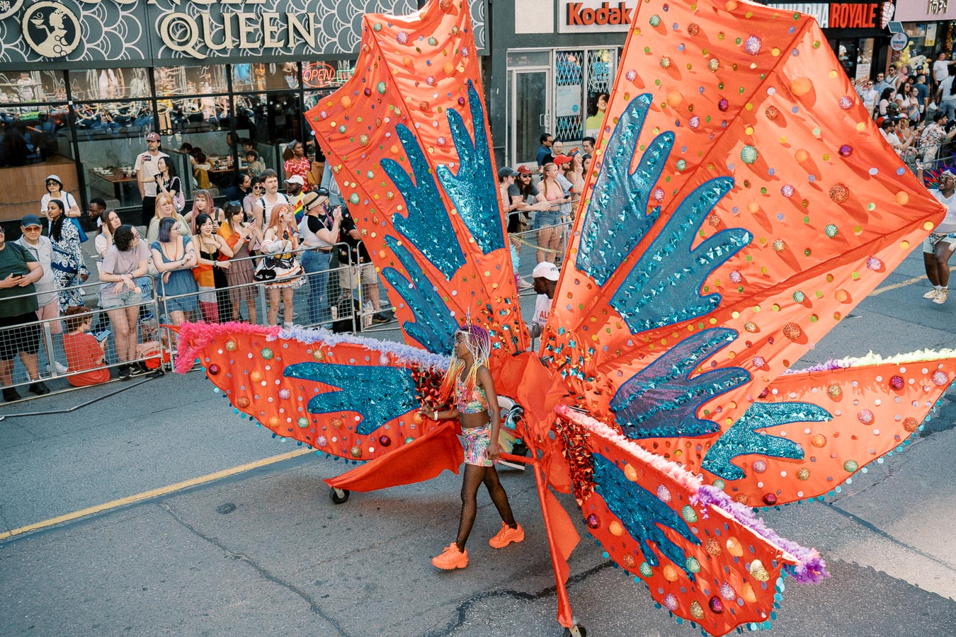 A vibrant parade participant wearing a large, colorful costume with red and blue wings captivates the crowd during a street festival.