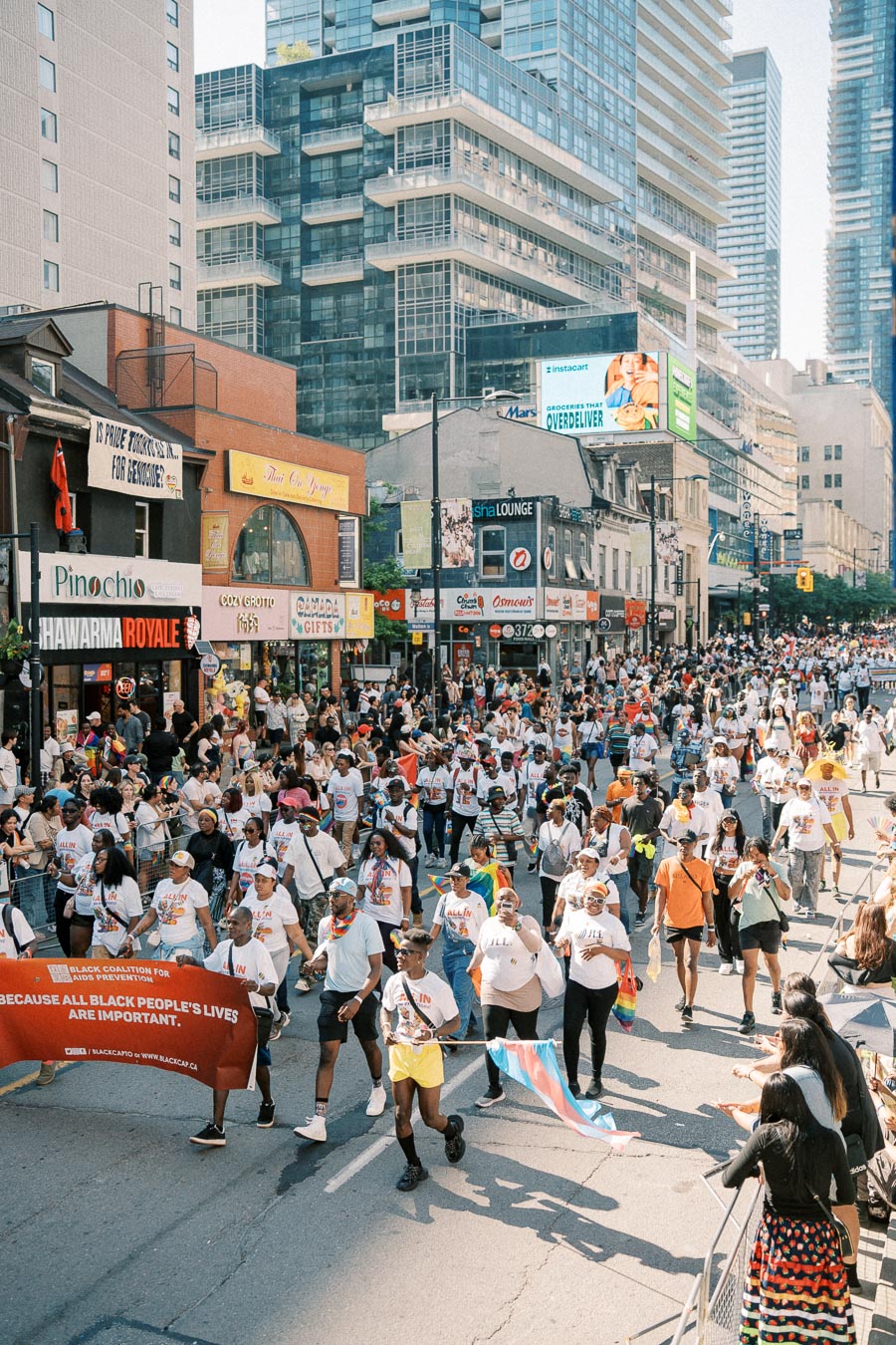 A vibrant parade takes place on a bustling city street, with participants holding banners and flags. The event is surrounded by tall buildings and local shops, creating a lively urban atmosphere.