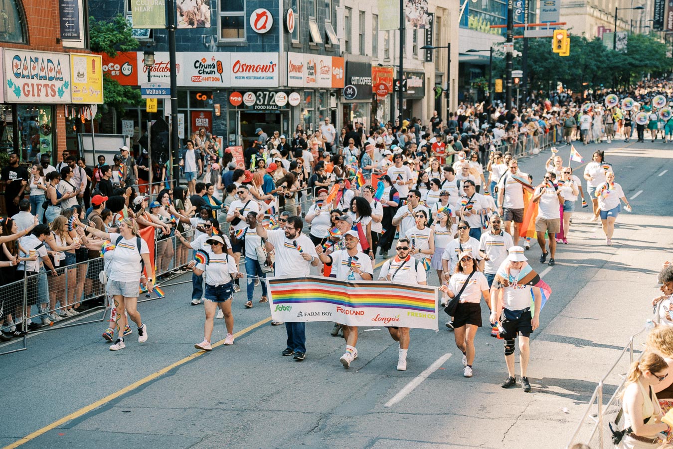 Participants in a pride parade march down a city street, holding a rainbow banner with company logos. Spectators cheer from the sidelines, celebrating diversity and inclusion.