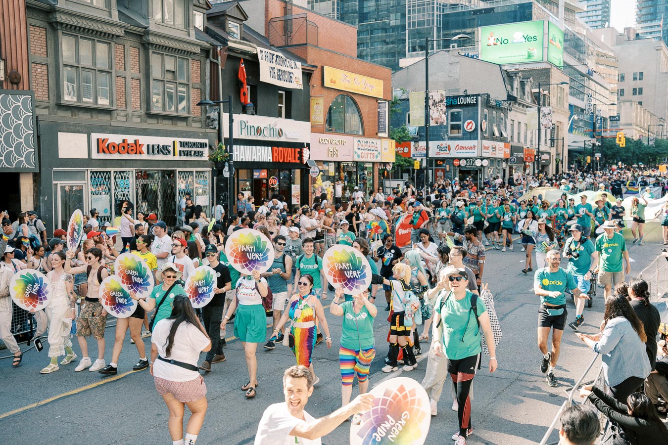 Participants in a vibrant street parade hold colorful signs and wear green shirts, celebrating amidst a crowd of spectators and various storefronts.
