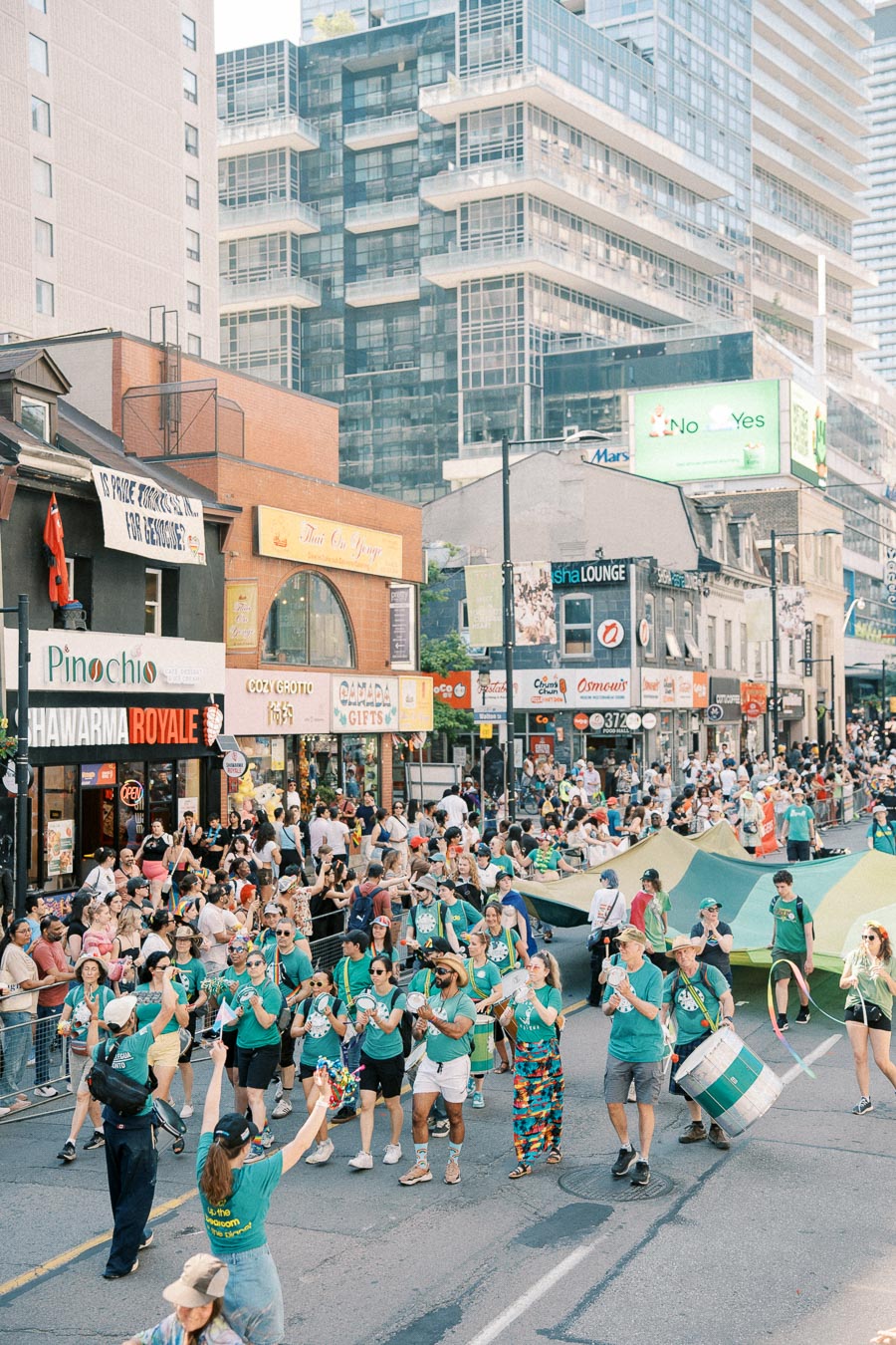 A lively parade with participants in green shirts marching down a bustling city street, surrounded by spectators and urban buildings.