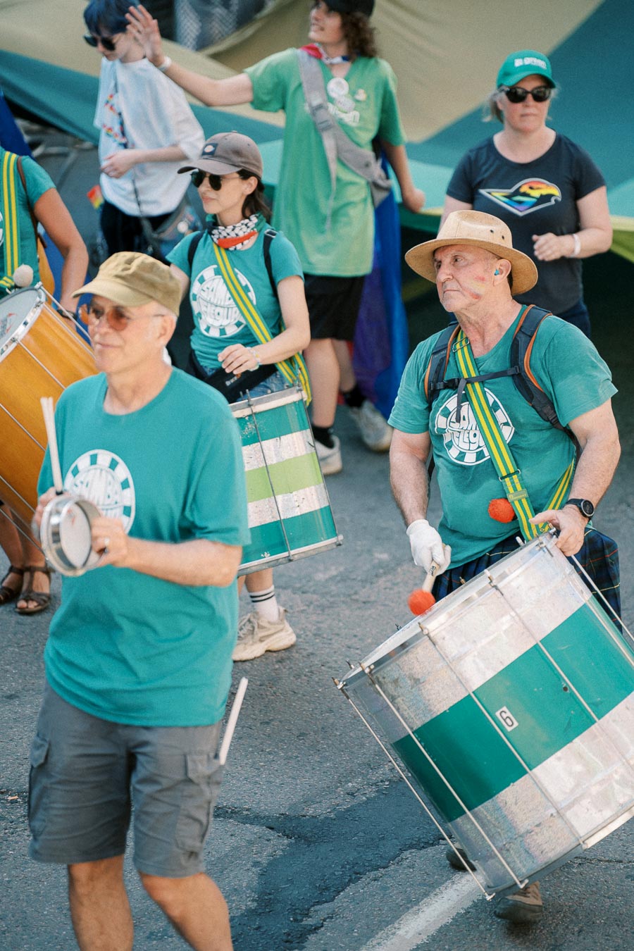 A group of people wearing green shirts participate in a parade, playing various drums. They appear to be part of a samba band, as indicated by the logos on their shirts. The scene is lively and festive, with participants focused on their performance.