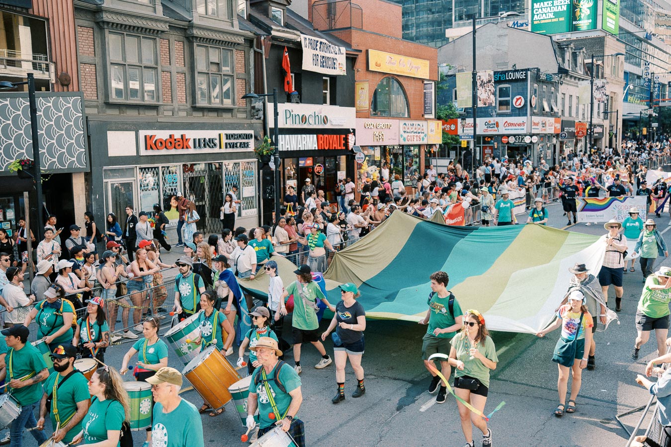 A vibrant parade scene with participants carrying a large green and yellow banner, accompanied by drummers, as spectators line the city street.