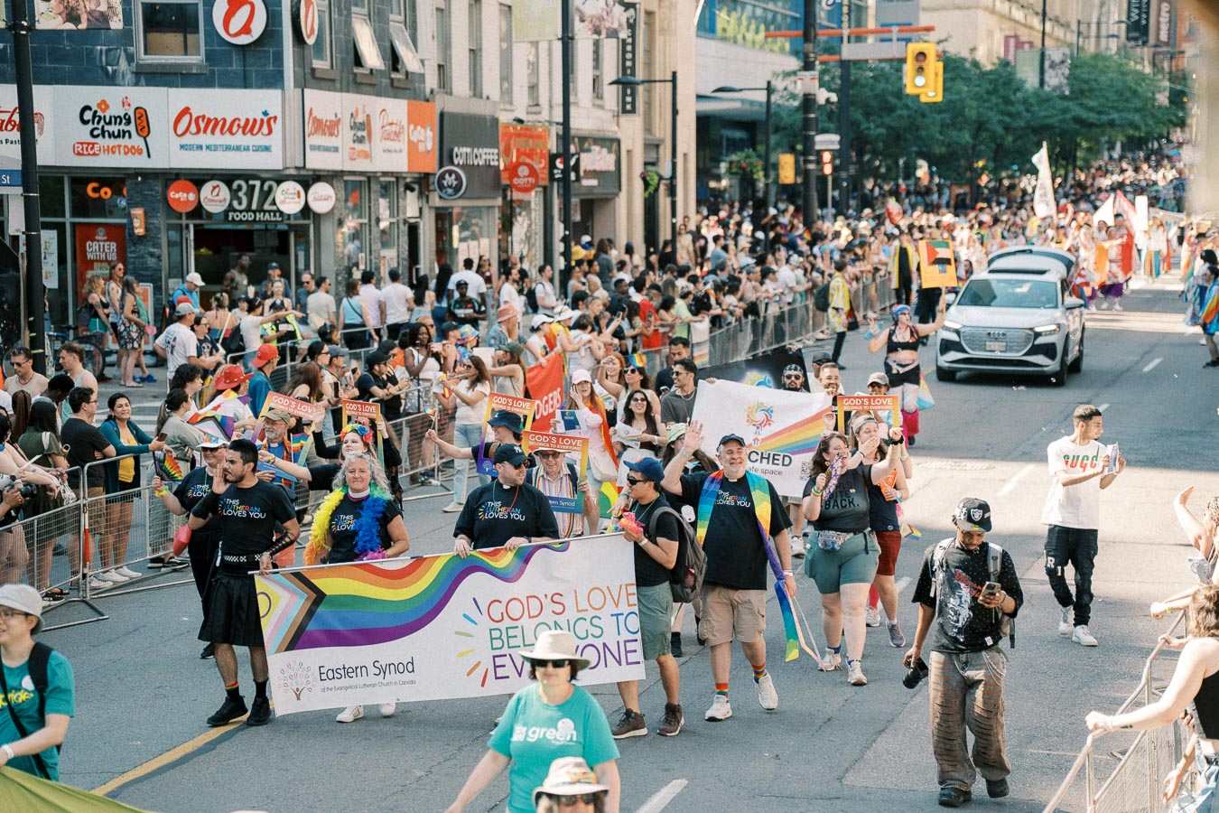 Participants march in a lively pride parade, holding banners promoting inclusivity and love, as a large crowd watches from the sidelines.