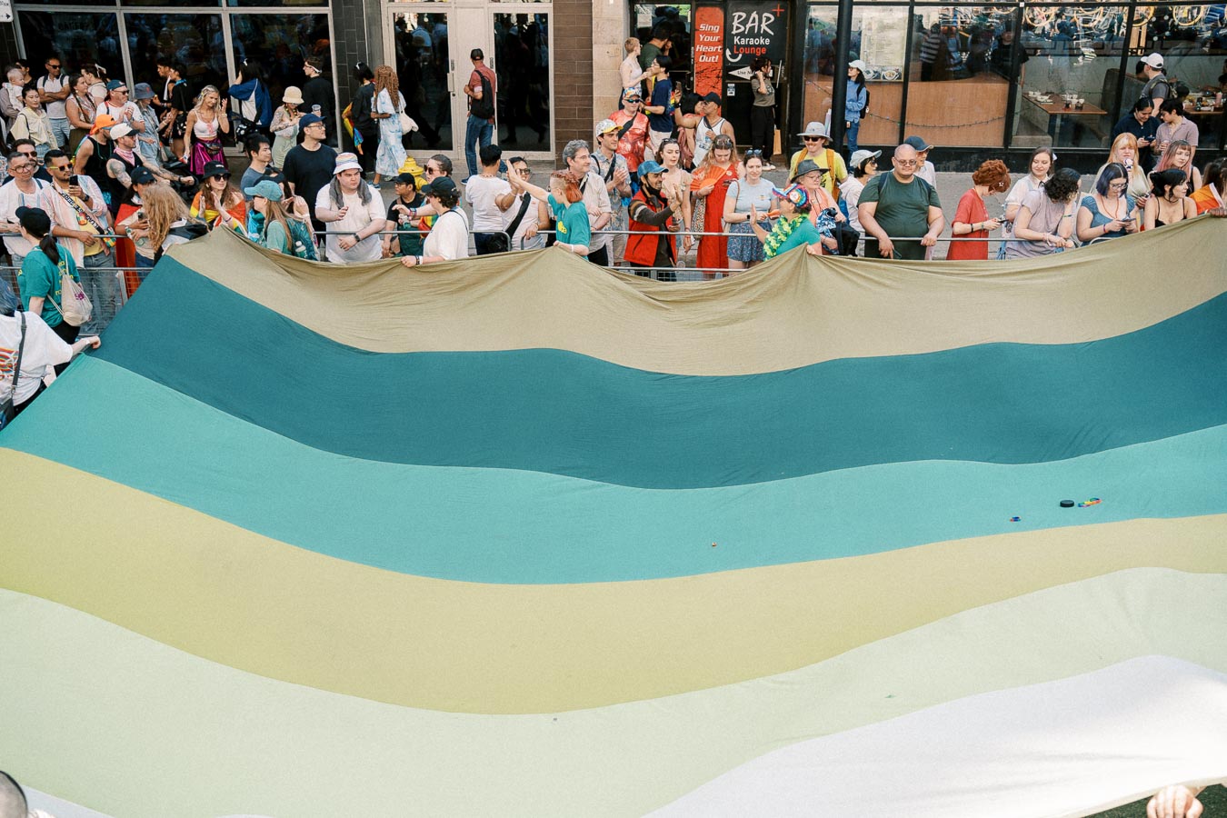 A vibrant crowd participates in a parade, holding a large flag with green and beige stripes.