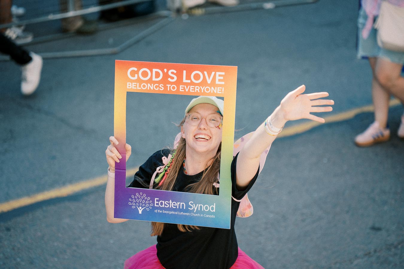 A person joyfully holds a colorful frame with the message 'GOD'S LOVE BELONGS TO EVERYONE!' during a public event. The frame also mentions the Eastern Synod of the Evangelical Lutheran Church in Canada.