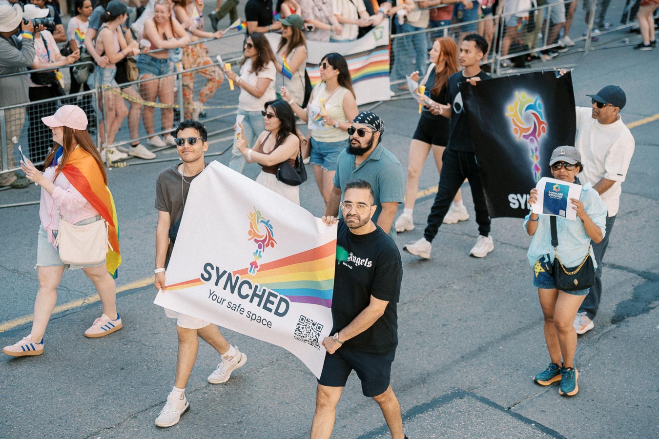 Participants in a pride parade hold banners with rainbow designs and the word 'SYNCHED', promoting inclusivity and safe spaces.