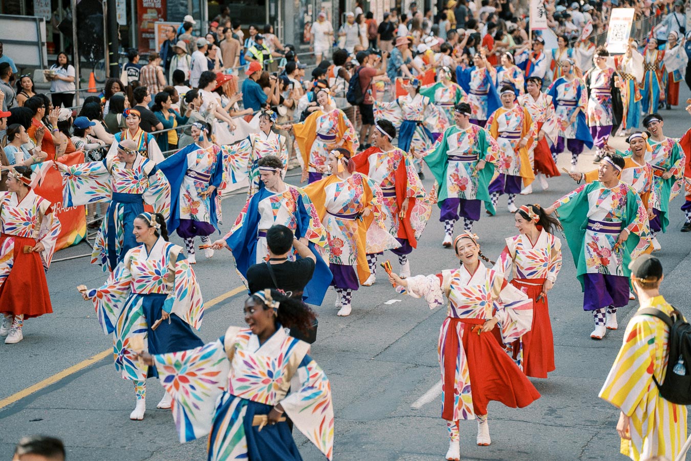 Participants in colorful traditional Japanese costumes dance joyfully during a vibrant street parade, surrounded by a large crowd of spectators.